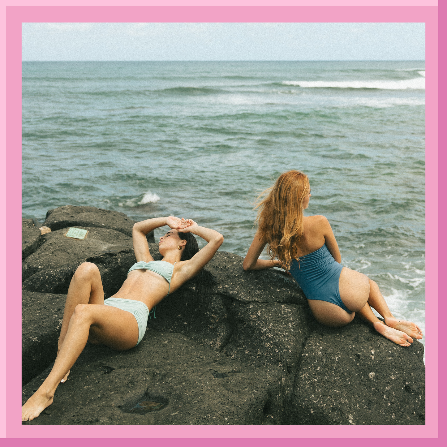 Two women in swimsuits sitting on rocks by the ocean.