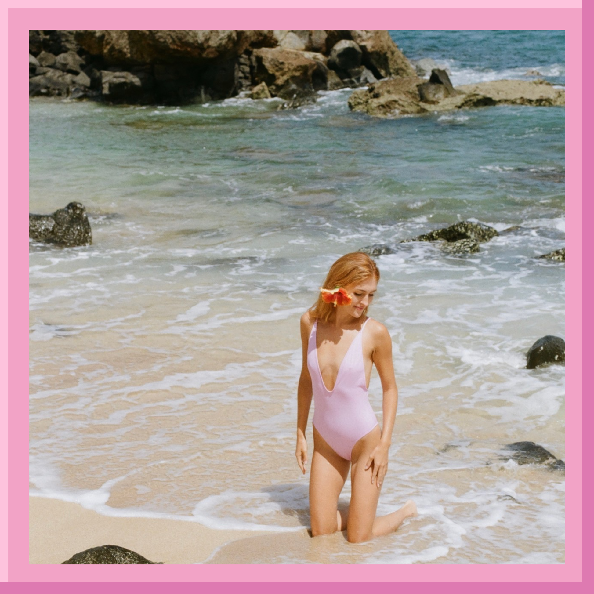 Woman in a pink swimsuit standing on a beach with rocks and water in the background.
