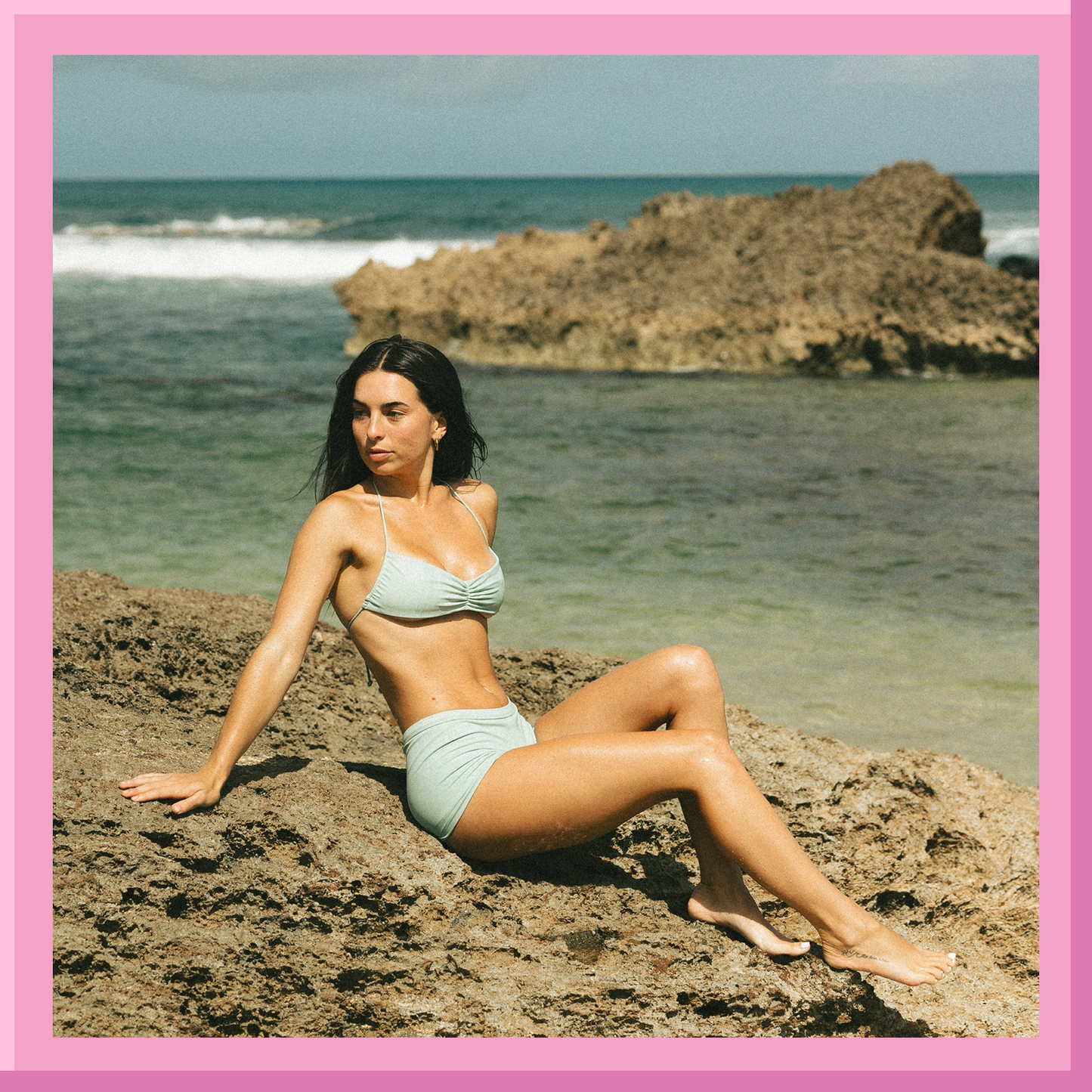 Woman in light blue bikini sitting on a rock by the ocean with a pink border.