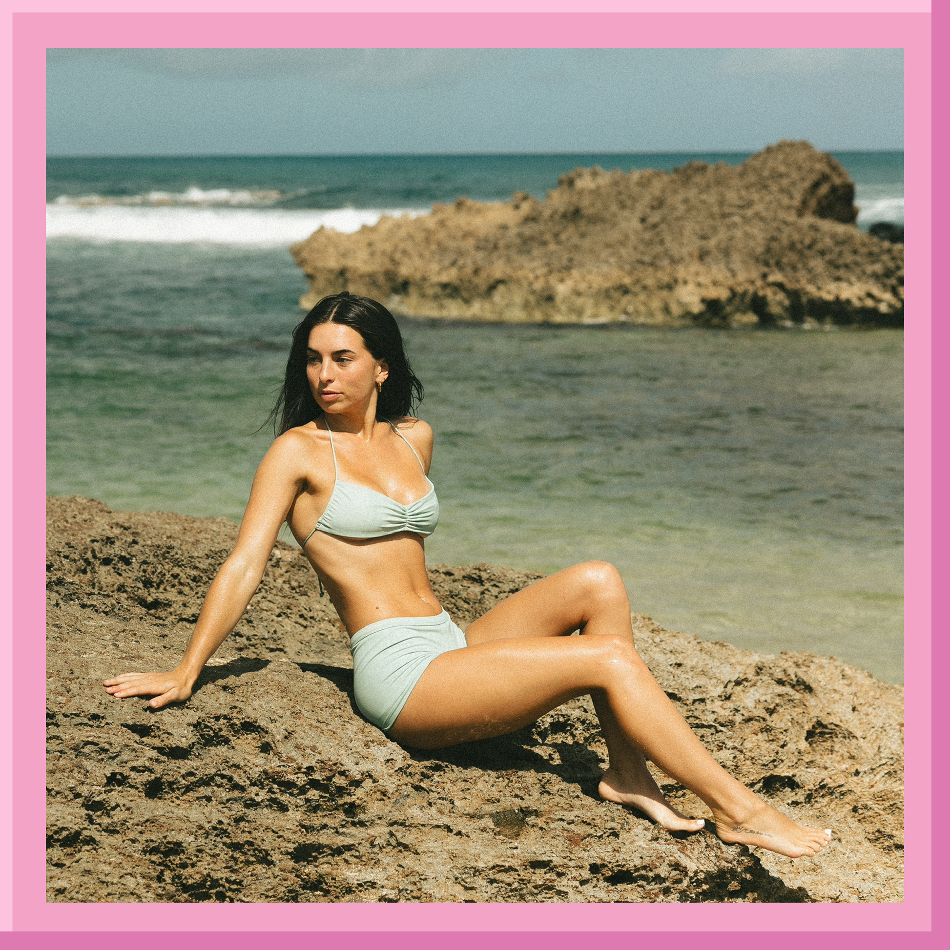 Woman in light blue bikini sitting on a rock by the ocean with a pink border.